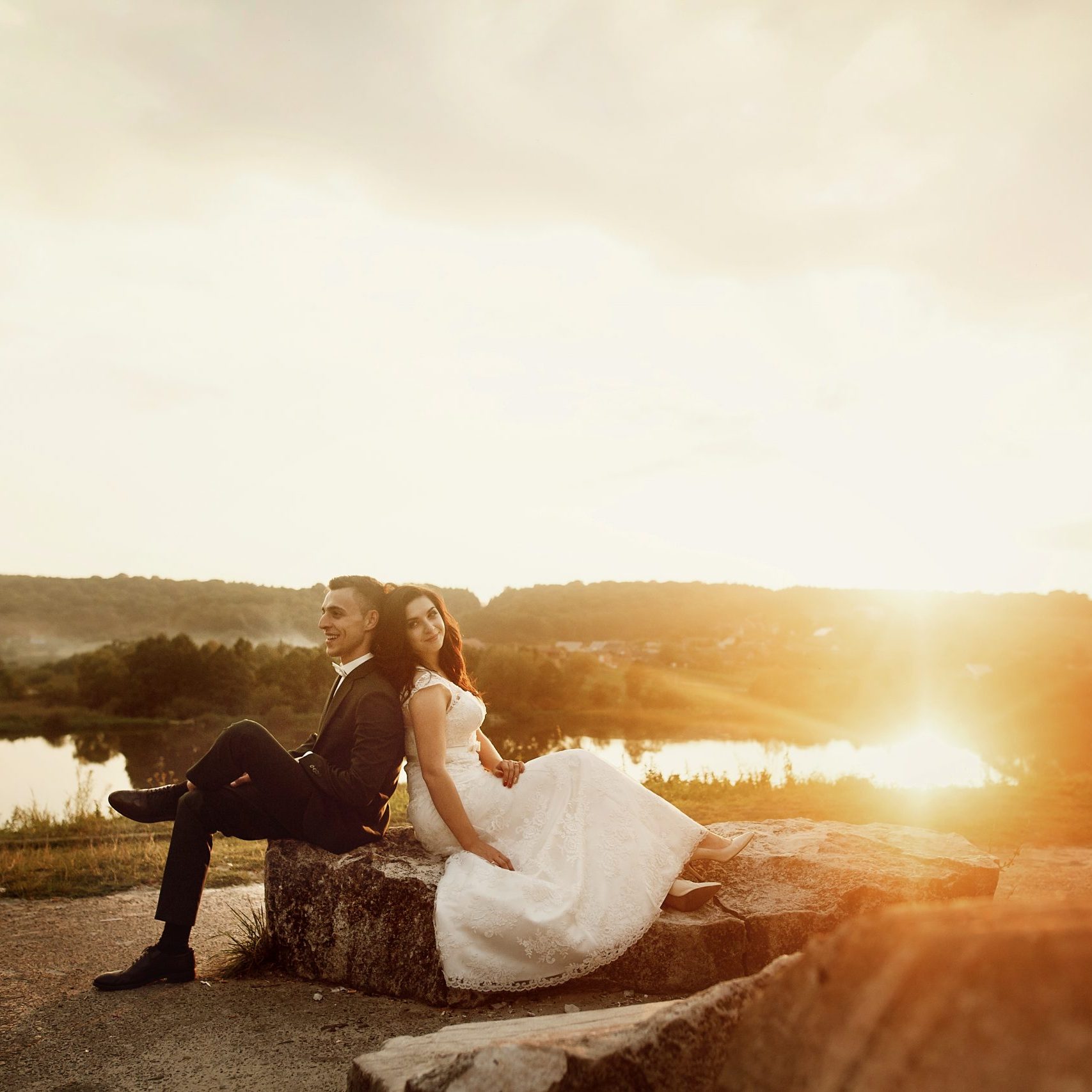 Romantic couple of newlyweds resting on stone bench at sunset field near lake landscape, happy bride leaning against smiling groom outdoors
