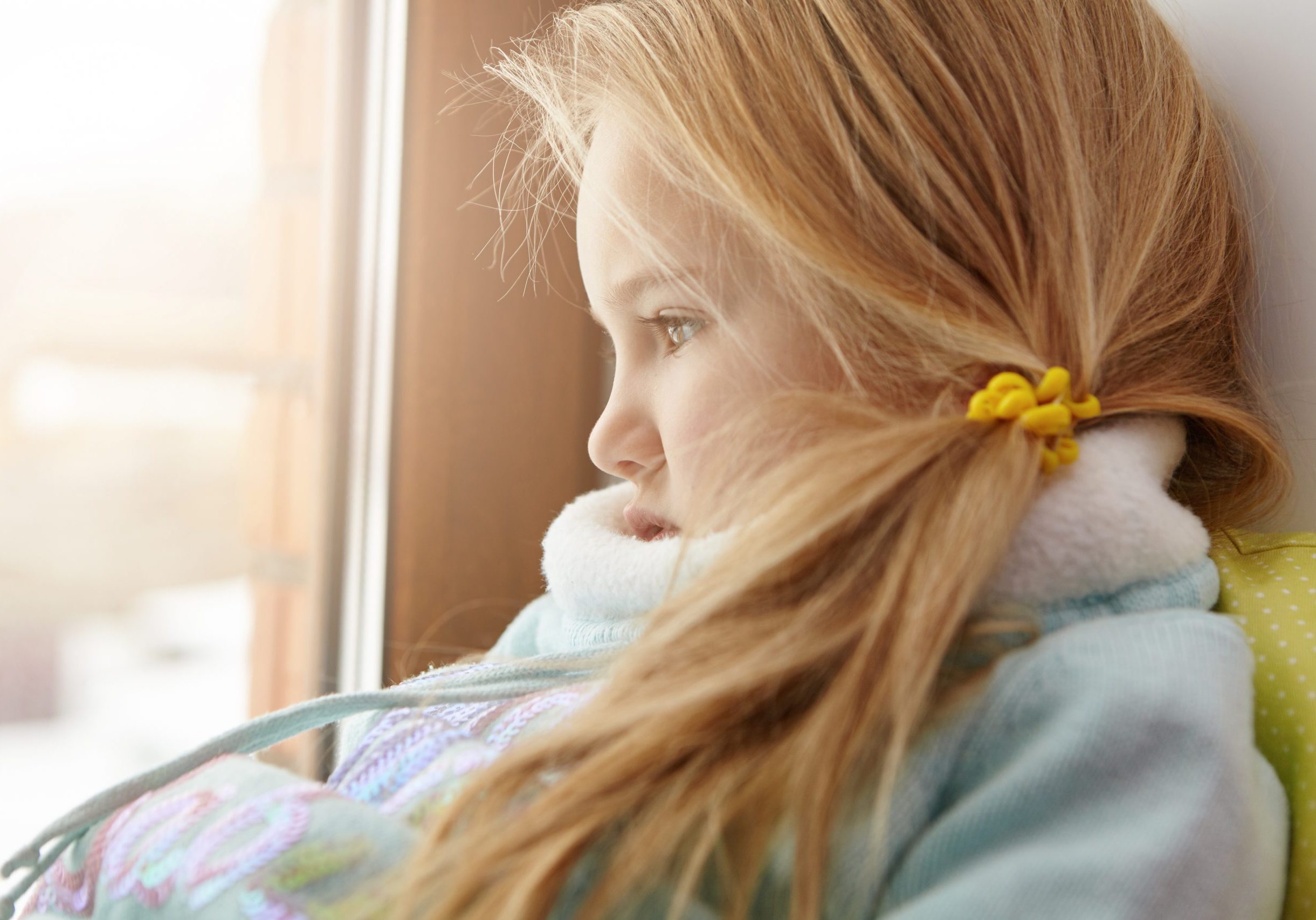 Sad little girl in warm clothes sitting alone on windowsill, looking out window, watching children play on street, forced to stay at home because of illness. Female child waiting for mother indoors